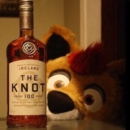 A bottle of 'The Knot' Irish whiskey, labeled 'Proof 100 Proof', sits on a surface next to a large, furry, cartoonish animal…