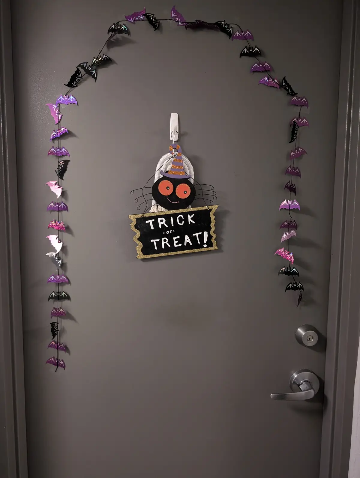A gray door is decorated for Halloween with a string of purple and black glittery bat cutouts forming an arch. Centered on…