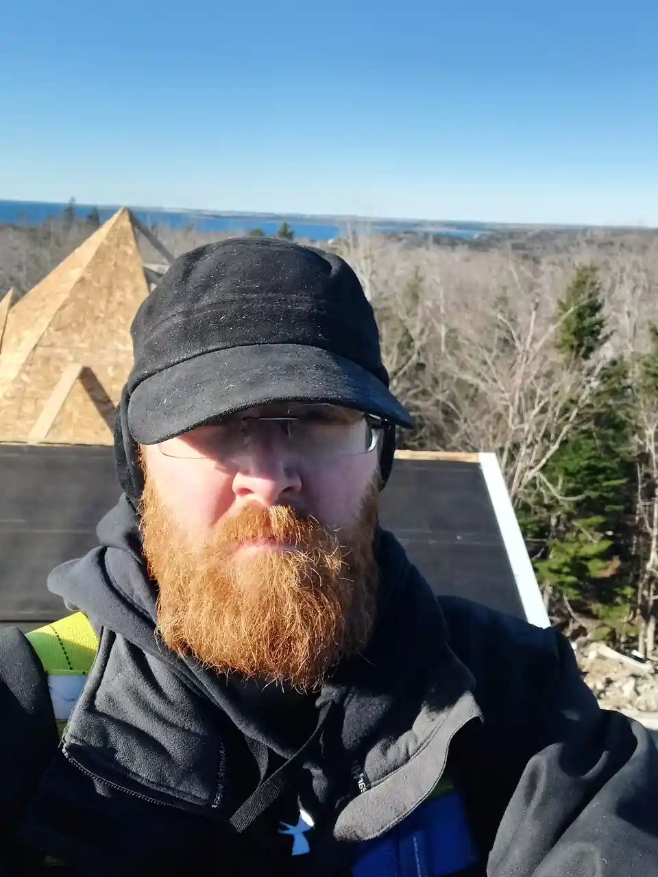 A man with a red beard, glasses, and a black cap takes a selfie on a construction site, standing on a roof with unfinished…