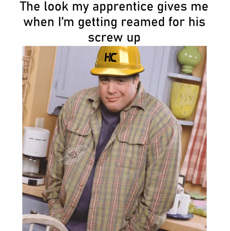 A man wearing a yellow hard hat with 'KC' on it and a plaid work shirt stands in a kitchen, smiling smugly with his hands in…