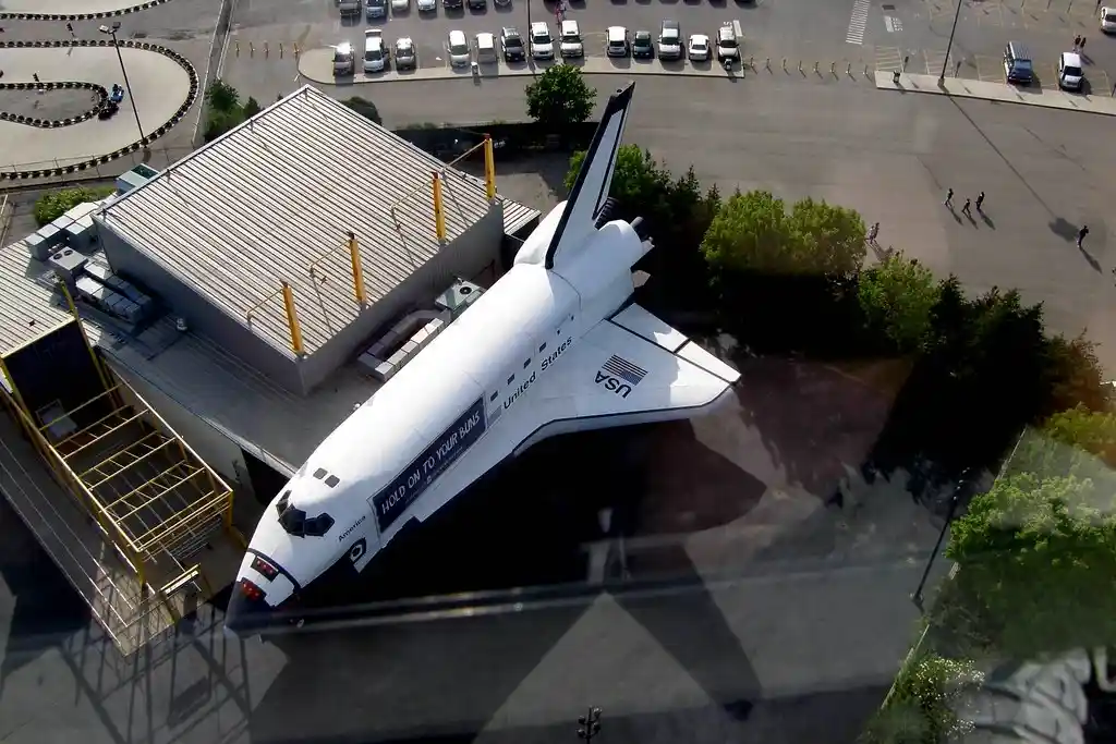 An aerial view shows a white space shuttle, labeled 'United States' and 'America', parked outside a building with a yellow…