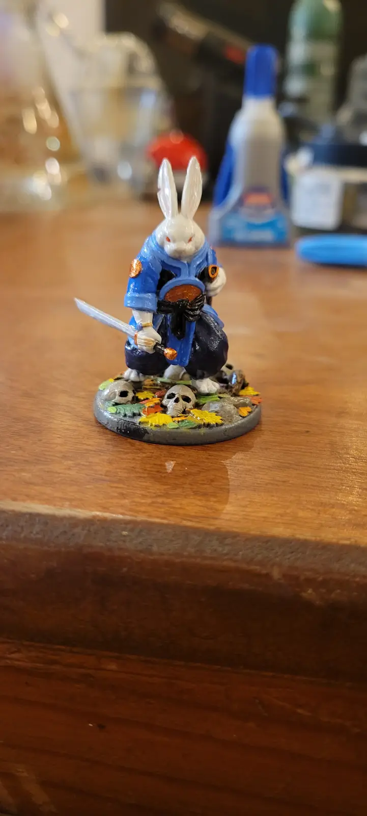 A painted miniature figurine of a white rabbit dressed in blue samurai armor, holding a sword and standing on a base with…
