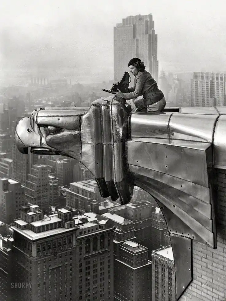 A black-and-white historical photograph shows a woman crouched atop a large, ornate metal gargoyle statue on a skyscraper…
