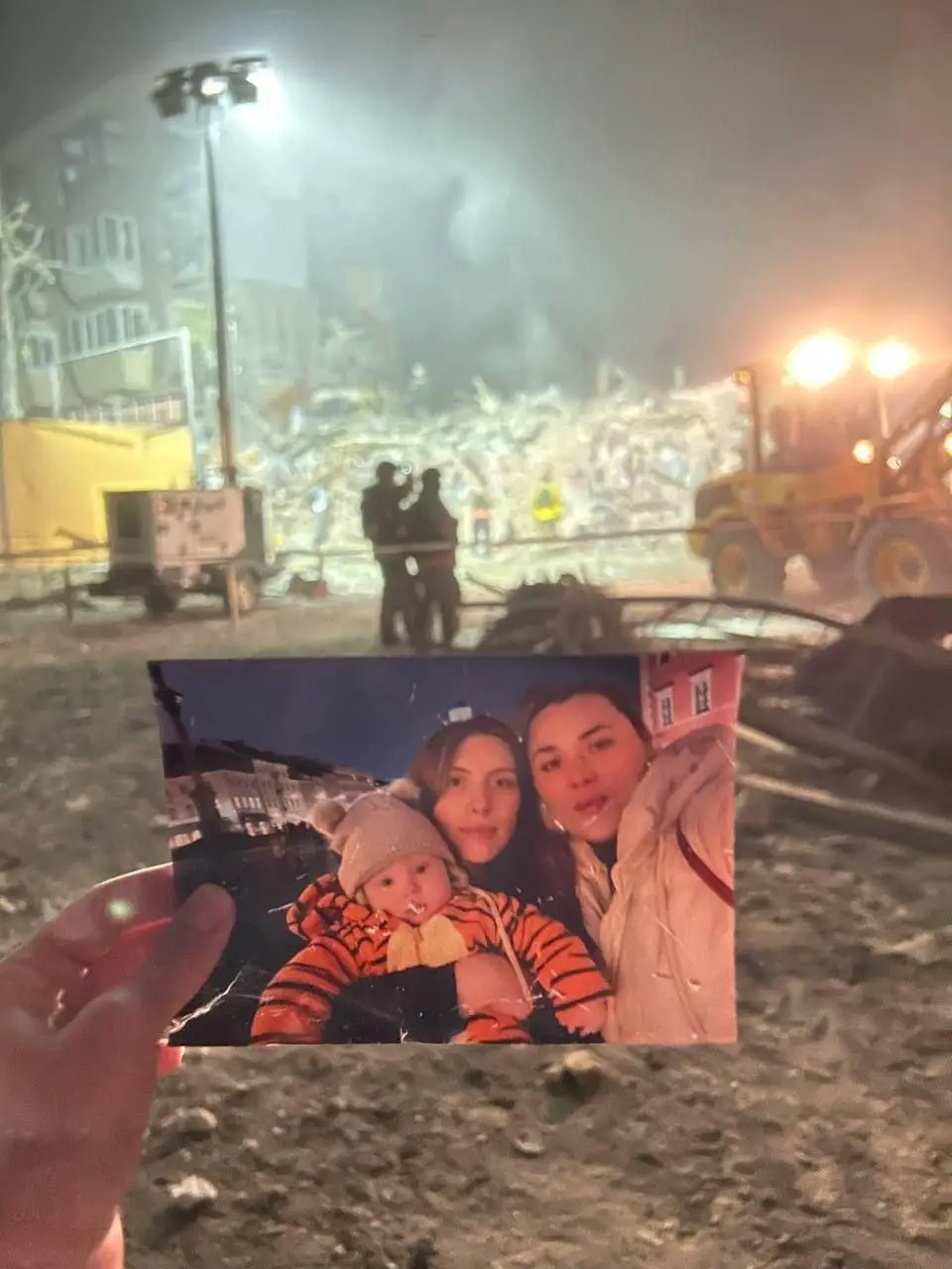 A hand holds up a photo of three people — two adults and a baby in a tiger-print outfit — smiling in front of a scenic city…