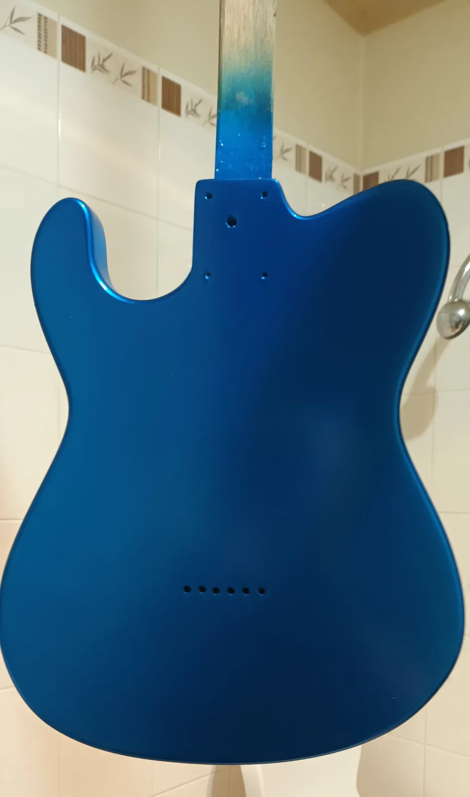 A blue electric guitar body is mounted vertically against a tiled bathroom wall, with a showerhead visible to the right. The…