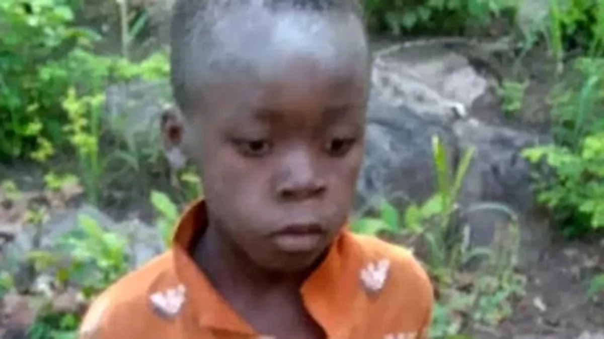 A young child with a solemn, downward-looking expression wears an orange shirt with white patches, standing outdoors amidst…