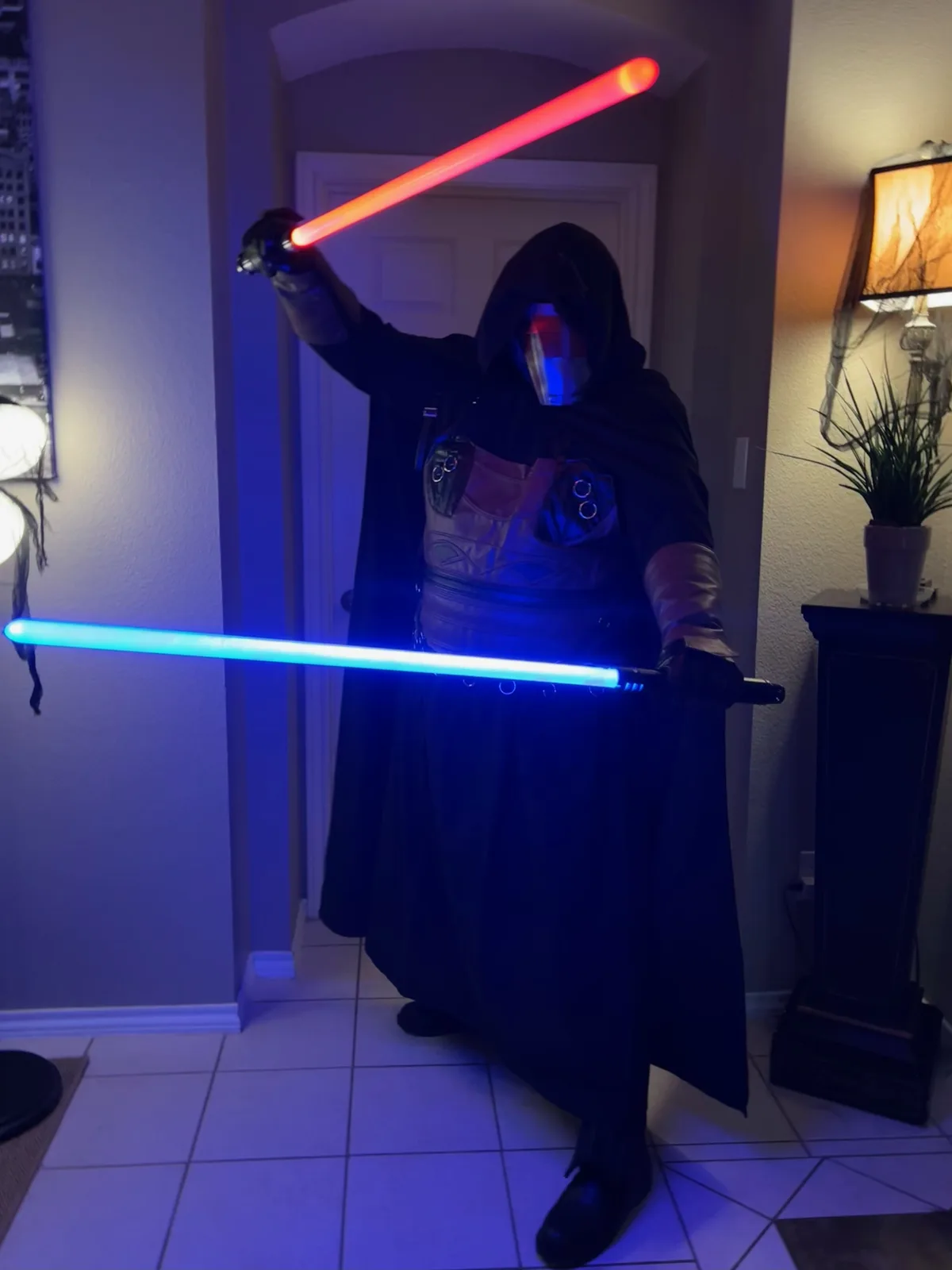 A person in a dark, hooded robe and mask stands indoors holding two glowing lightsabers, one red in their raised right hand…