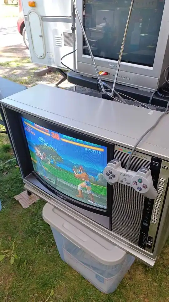 An old silver Sony CRT television sits outdoors on a plastic storage bin, displaying a colorful 2D fighting game with a…