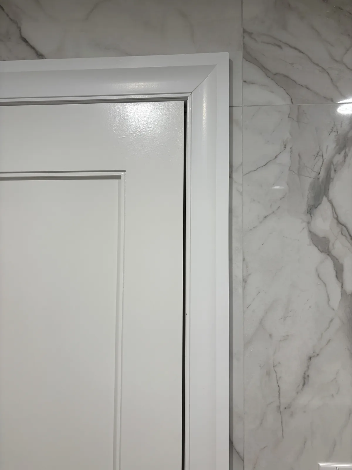 A close-up photo shows a white interior door slightly ajar, revealing a narrow black gap between the door and its white…