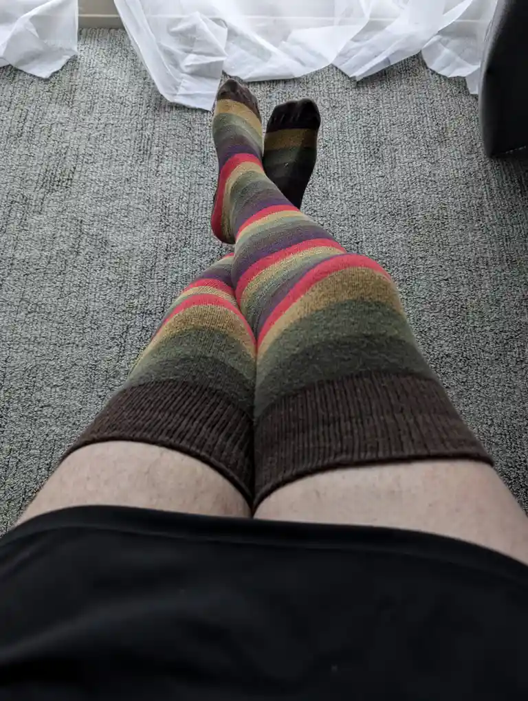 A first-person view of someone's legs wearing colorful striped knee-high socks, sitting on a gray carpet with white plastic…