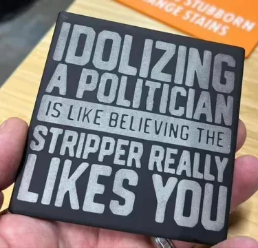 A hand holds a black square coaster with silver text that reads 'IDOLIZING A POLITICIAN IS LIKE BELIEVING THE STRIPPER…