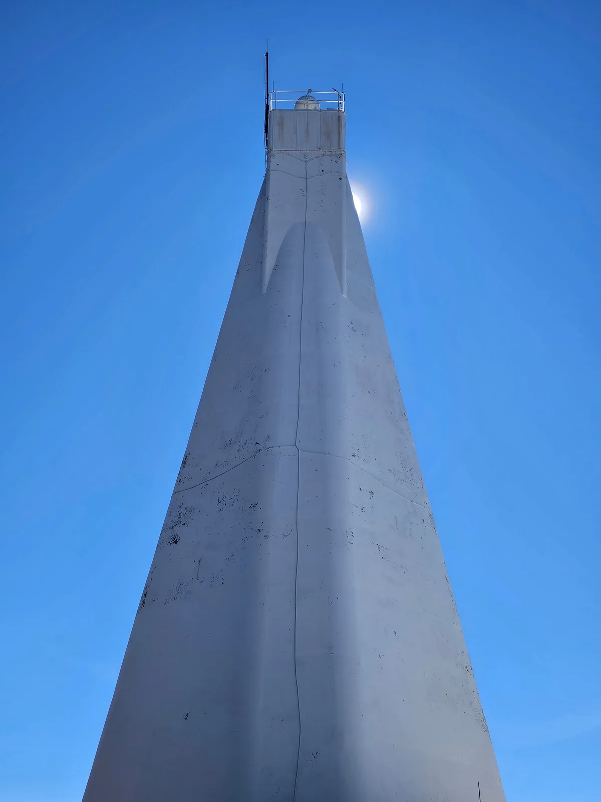 A low-angle shot of a tall, white, conical lighthouse against a clear blue sky, with the bright moon visible behind its…