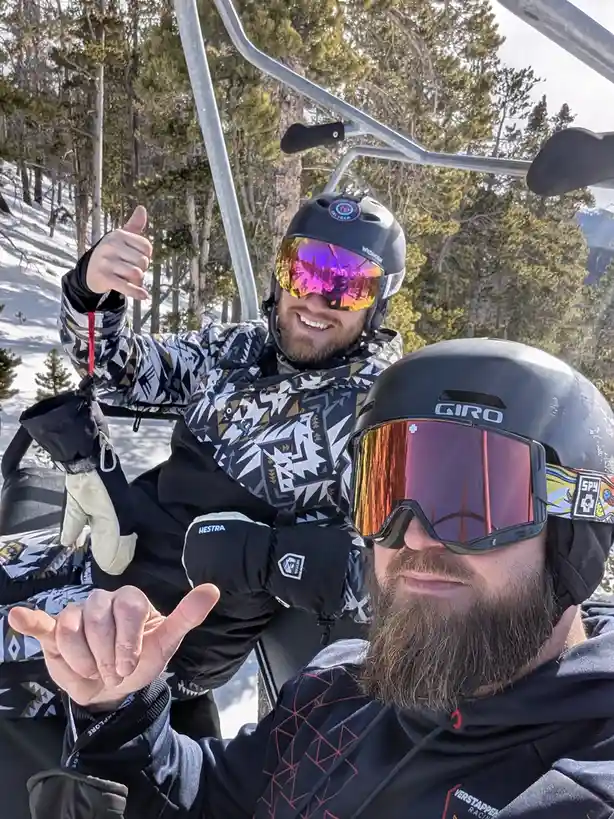 Two men in ski gear on a chairlift, both making the shaka hand gesture. The man in the foreground has a beard and wears a…