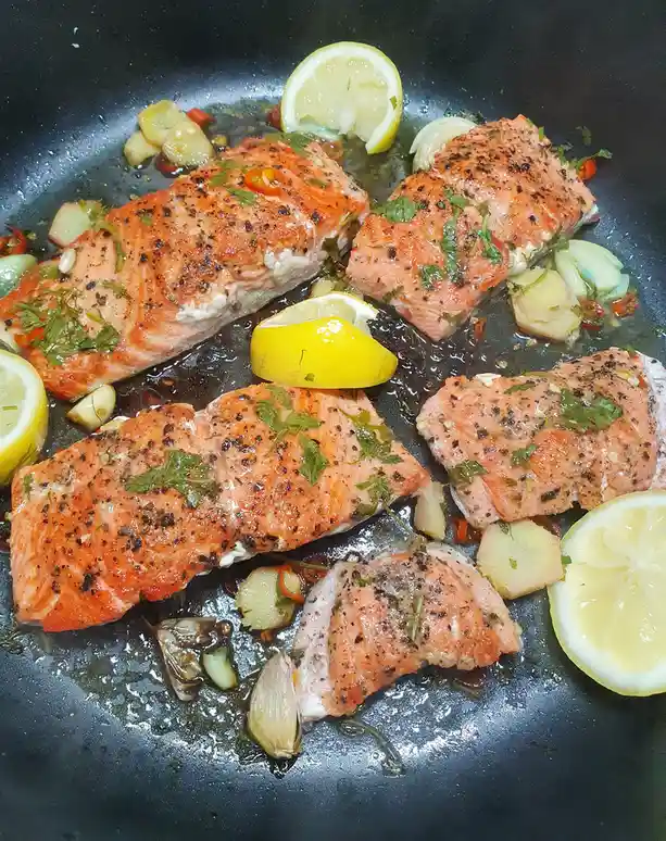 A close-up photo shows four pieces of cooked salmon fillets sizzling in a black frying pan, garnished with lemon wedges…