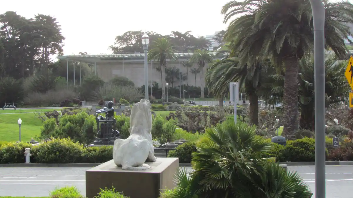 A photograph of a white marble lion statue sitting on a pedestal in a landscaped park with palm trees and greenery. The…