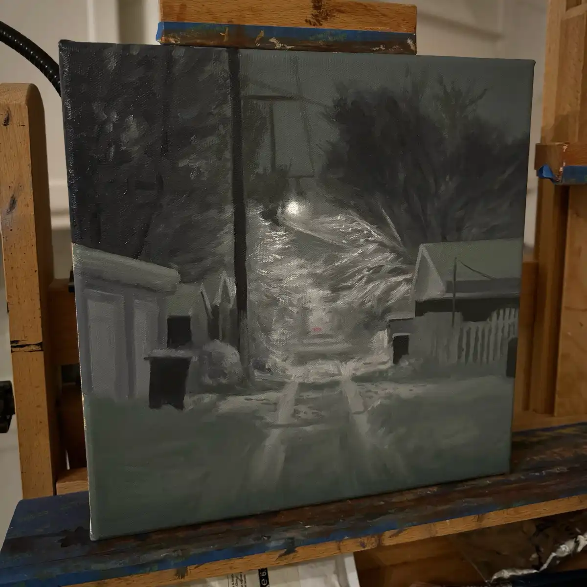 A painting of a snowy street scene at night, rendered in mostly gray and white tones, sits on an easel. The composition…