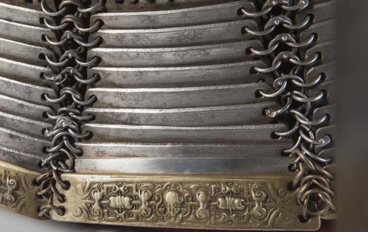 A close-up of ornate, metallic armor featuring horizontal plates and chainmail lacing, with a decorative brass band at the…