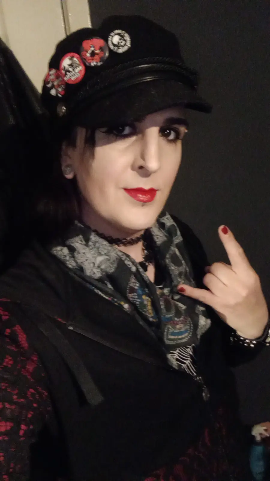 A person with dark makeup, red lipstick, and a black cap adorned with multiple skull and band-themed pins poses directly at…