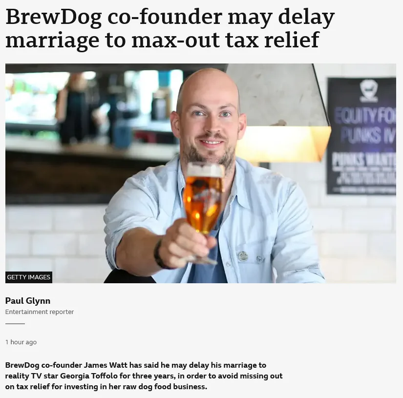 A bald man with a beard, wearing a light blue shirt, smiles while holding a glass of beer toward the camera. The image is…