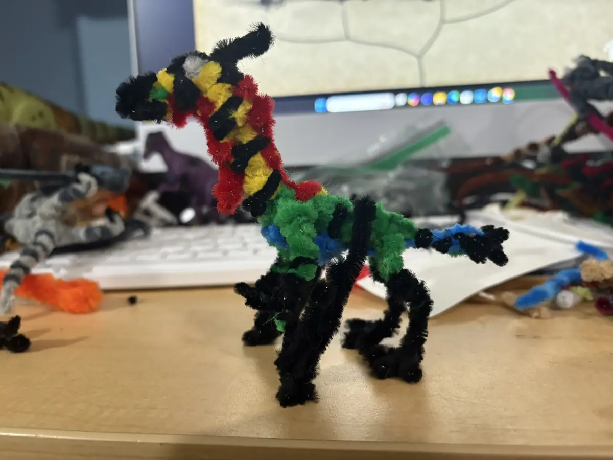 A colorful, crudely crafted dinosaur made from pipe cleaners stands on a wooden desk in front of a computer monitor. The…