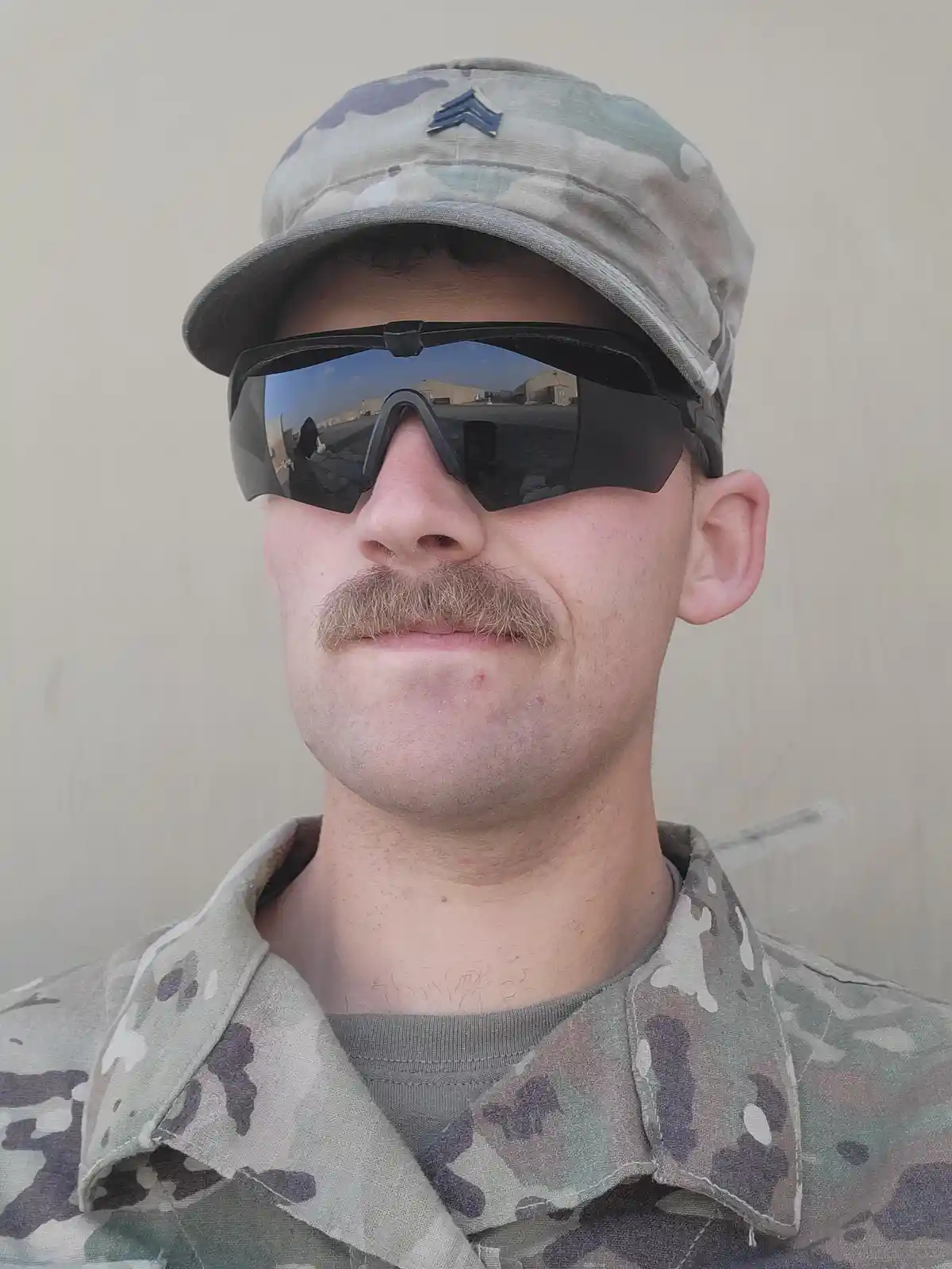 A man in military camouflage uniform and cap, wearing large, dark, wraparound sunglasses that obscure his eyes, has a…