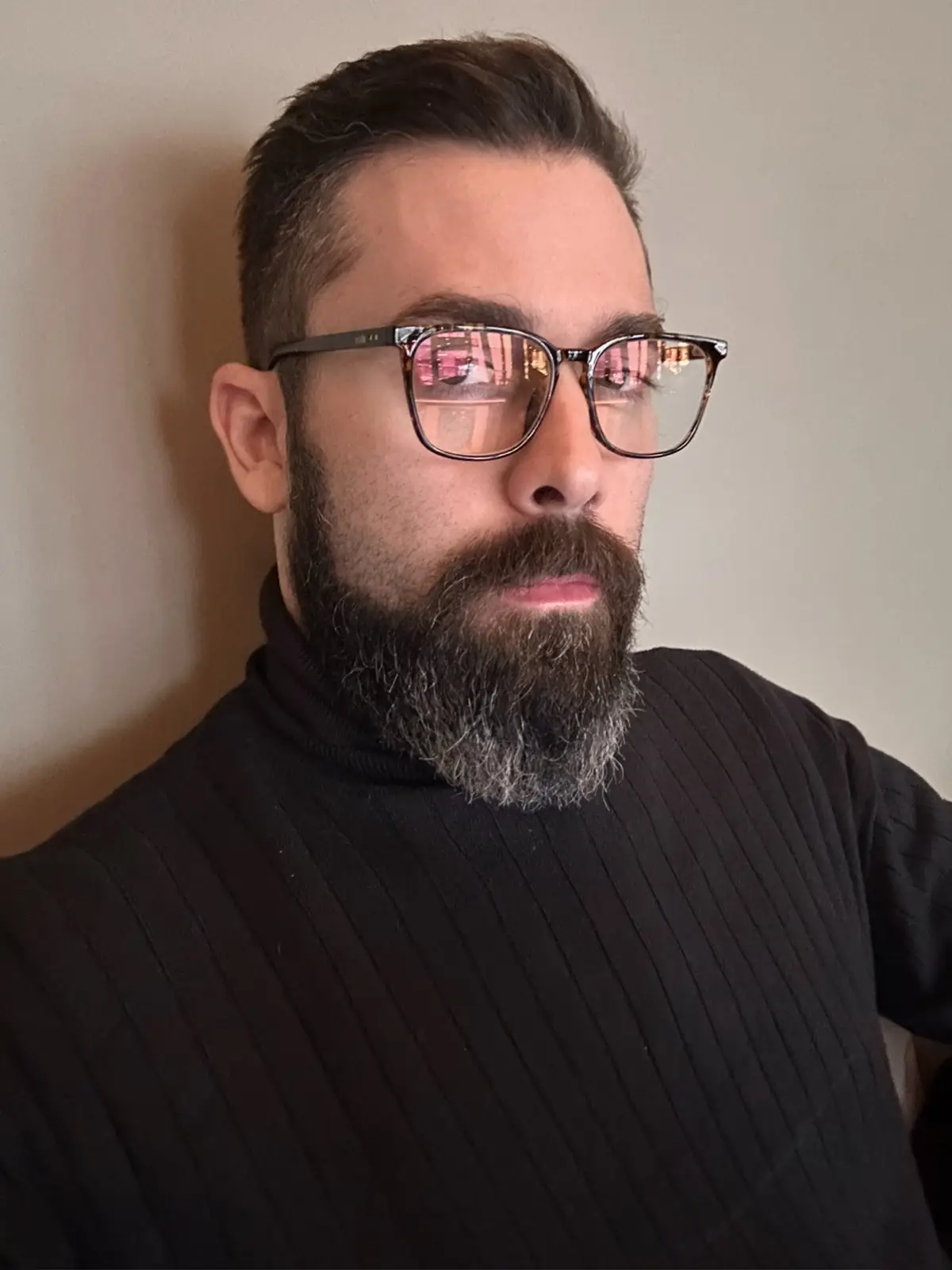 A man with a full beard and glasses, wearing a black ribbed turtleneck, looks off to the side with a neutral expression. The…