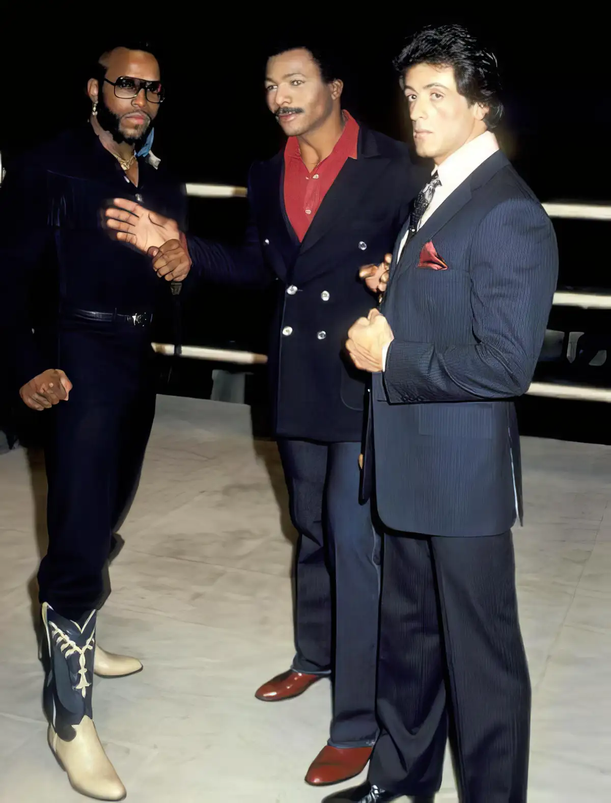 Three men in 1980s-style suits stand together, one with cowboy boots and sunglasses, another with a mustache and red shirt…