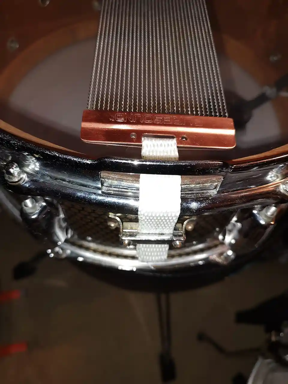 A close-up photo of a snare drum with a copper-colored 'PURESOUND' brand resonator attached to its head, secured by a white…