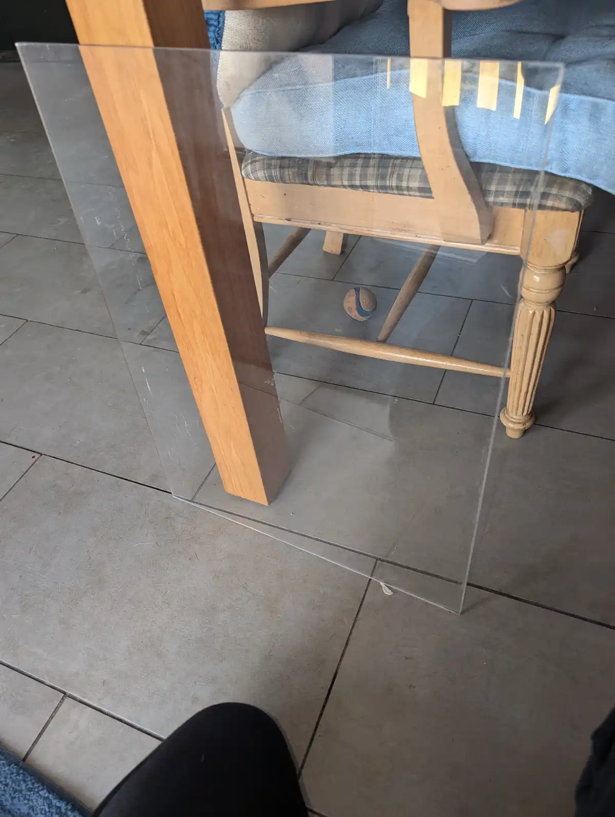 A low-angle photo shows a clear glass table with wooden legs, a wooden chair with a blue cushion and plaid seat, and a small…