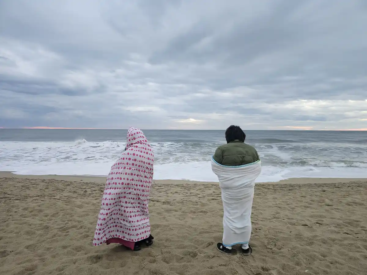 Two people stand on a sandy beach facing the ocean under a cloudy sky, both wrapped in blankets like wearable coats. One…