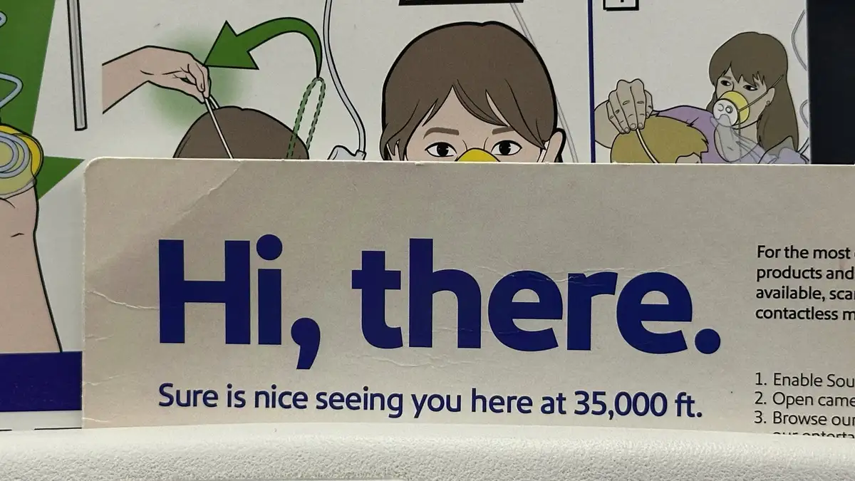 A close-up of an airplane safety card showing cartoon illustrations of passengers using oxygen masks. The card's main text…