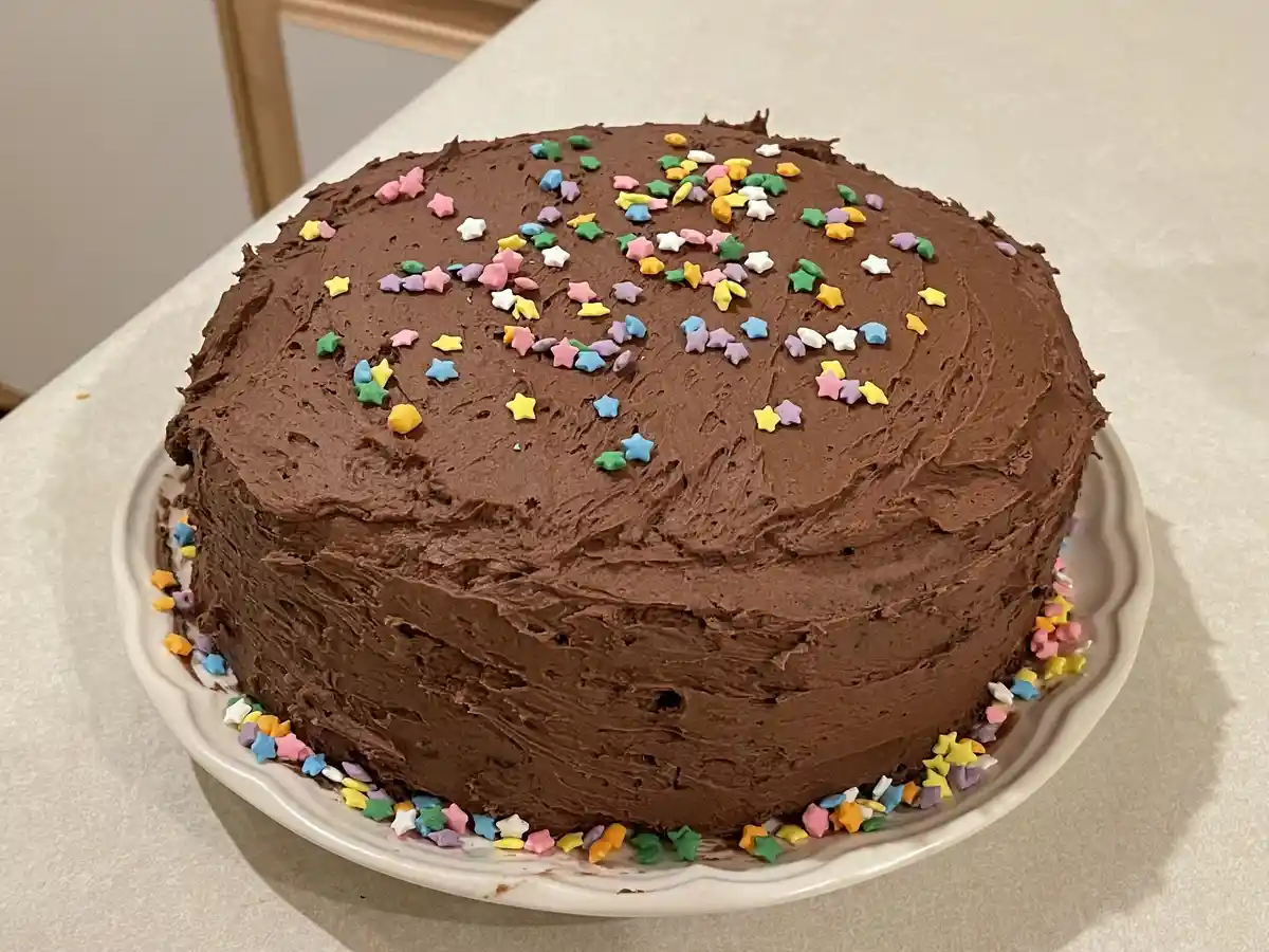 A round chocolate cake with thick frosting sits on a white plate, decorated with colorful star-shaped sprinkles scattered…