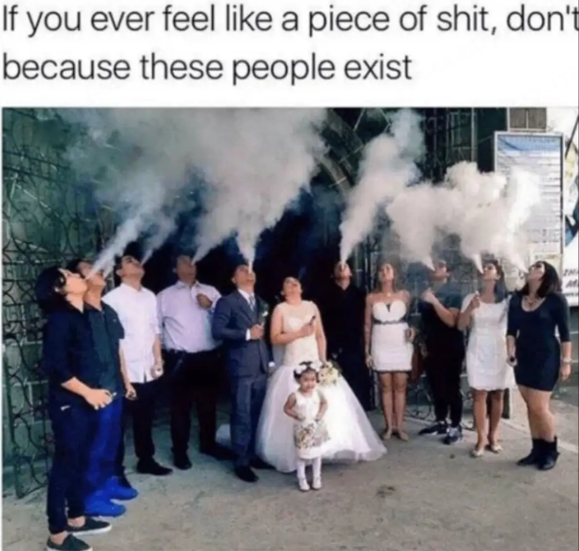 A group of people, including a bride and groom, are standing outdoors and exhaling large clouds of vapor from their mouths…