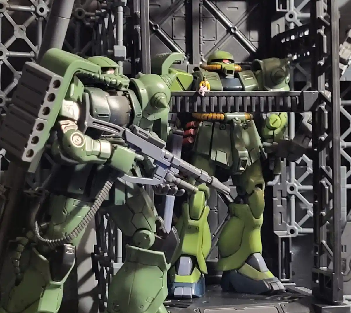 Two green, detailed mecha model figures stand in a metallic, industrial-style display rack. The foreground mecha holds a…