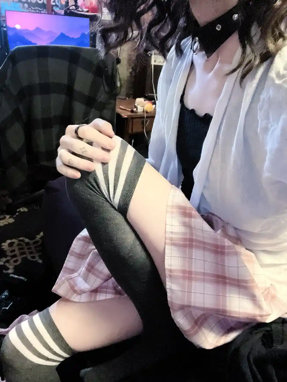 A person with dark curly hair is sitting in a chair, wearing a white sheer cardigan over a black top, a pink plaid skirt…