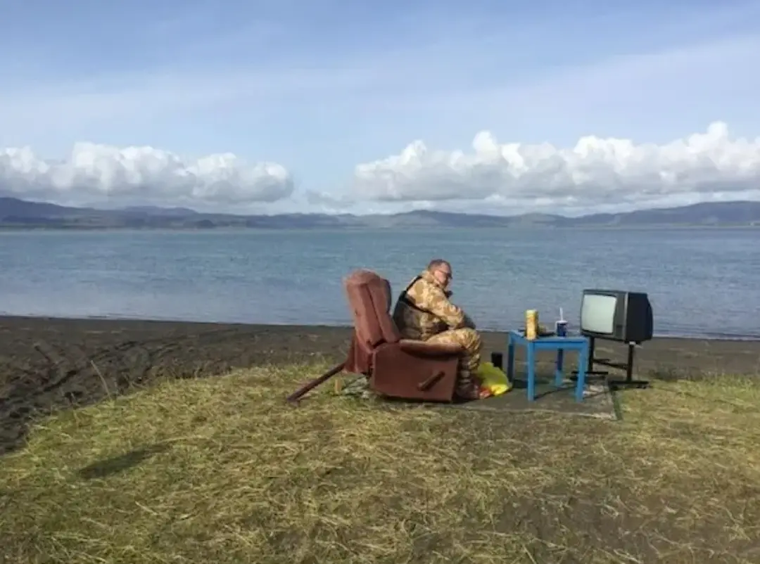 A man in camouflage military uniform sits in a brown recliner on a grassy beach next to a large body of water, watching an…