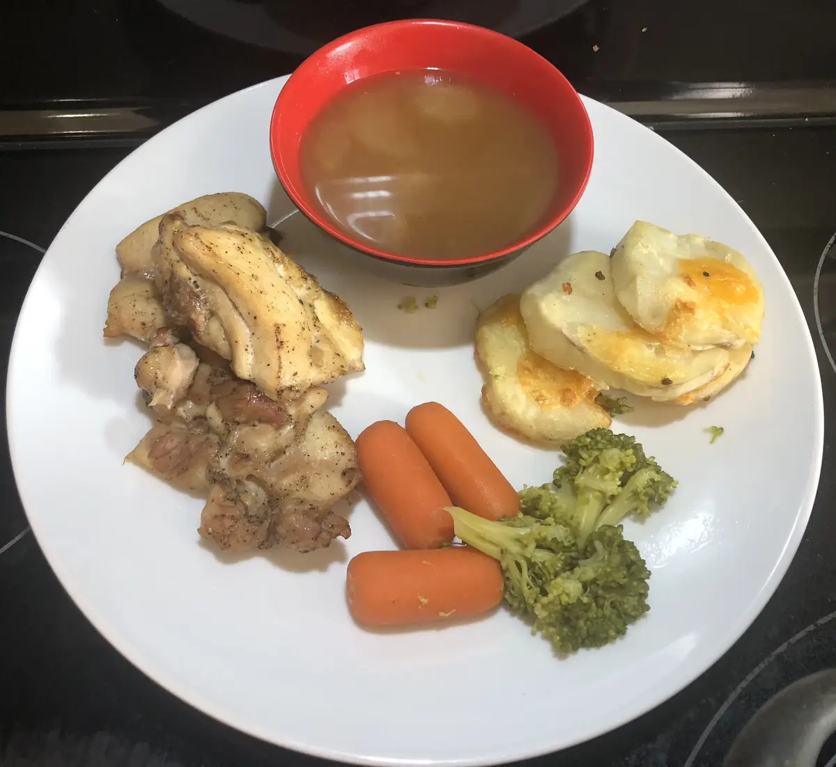 A white plate holds a meal of cooked chicken pieces, three baby carrots, a small floret of broccoli, and three slices of…