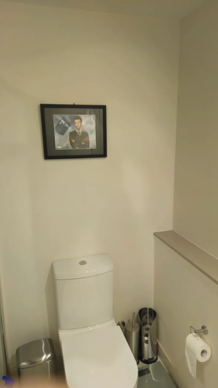 A bathroom with a white toilet, a framed signed photo of David Tennant as the Doctor from Doctor Who hanging above it, a…