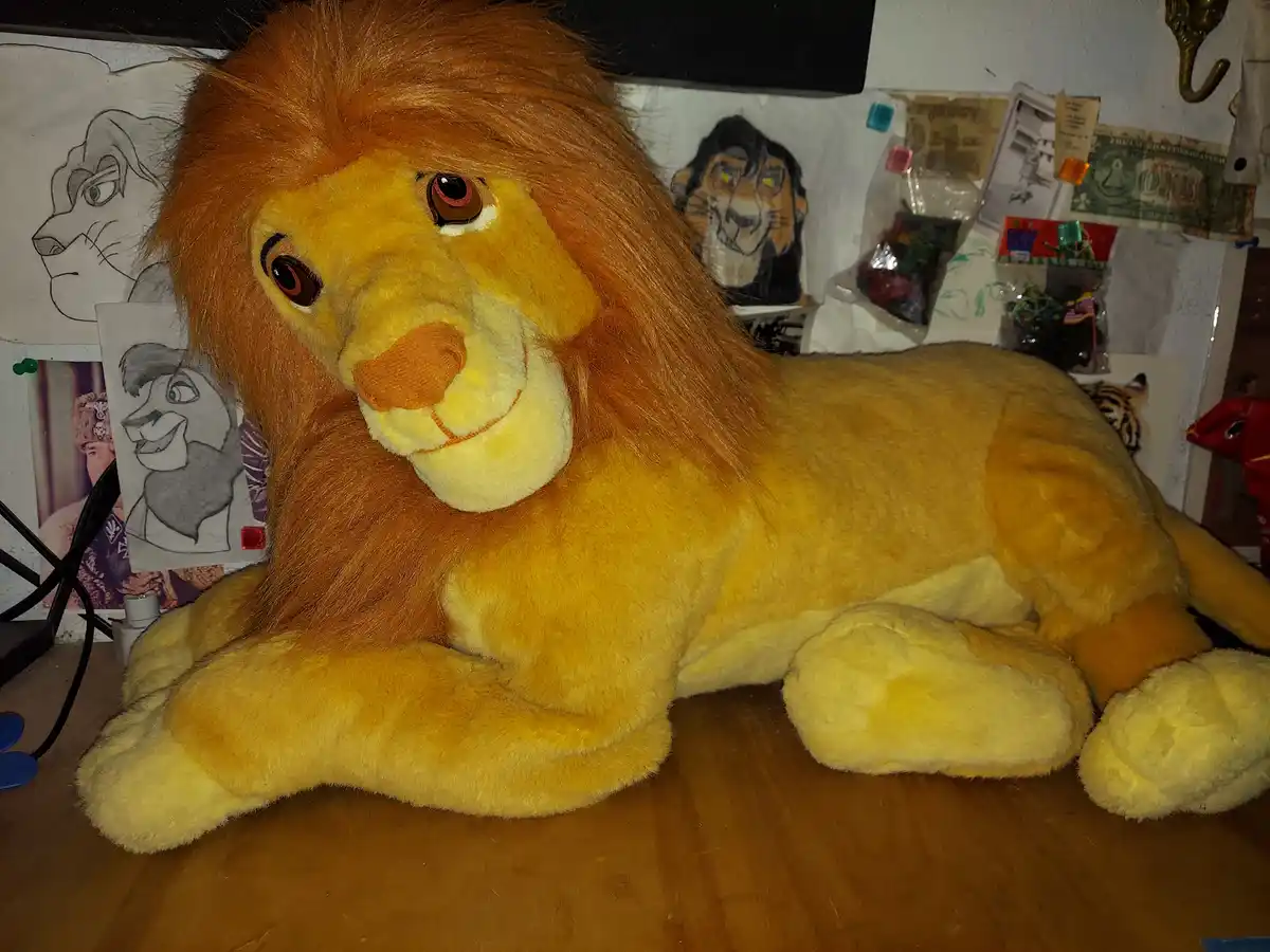A large, yellow plush lion toy with a full mane is lying on a wooden surface, looking directly at the camera with a gentle…