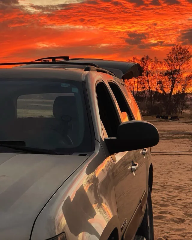 A silver SUV is parked on sandy ground under a vibrant orange and red sunset sky, with trees visible in the background. The…