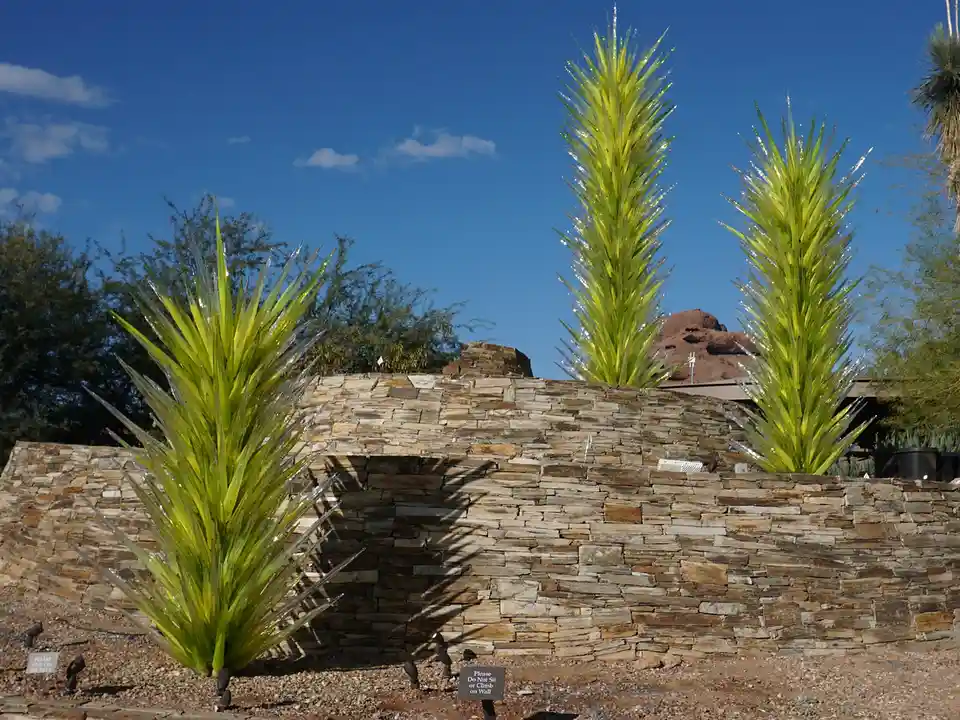 Three large, spiky, glass-like sculptures resembling yucca plants are planted in front of a low stone wall under a clear…