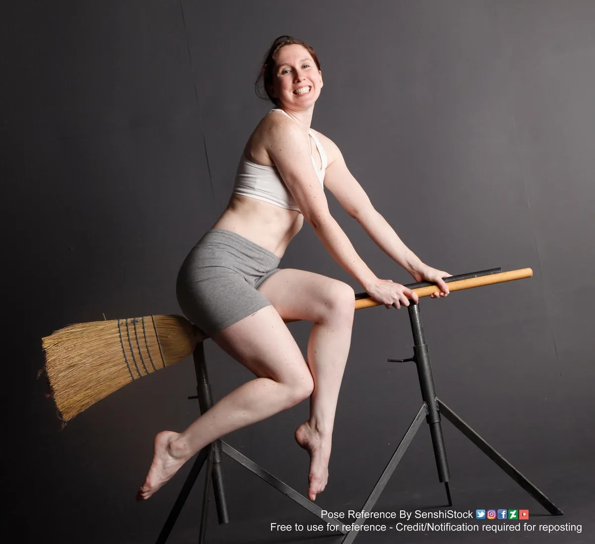 A woman in a white sports bra and grey shorts is posing with a broom, sitting on a black tripod stand with her legs dangling…