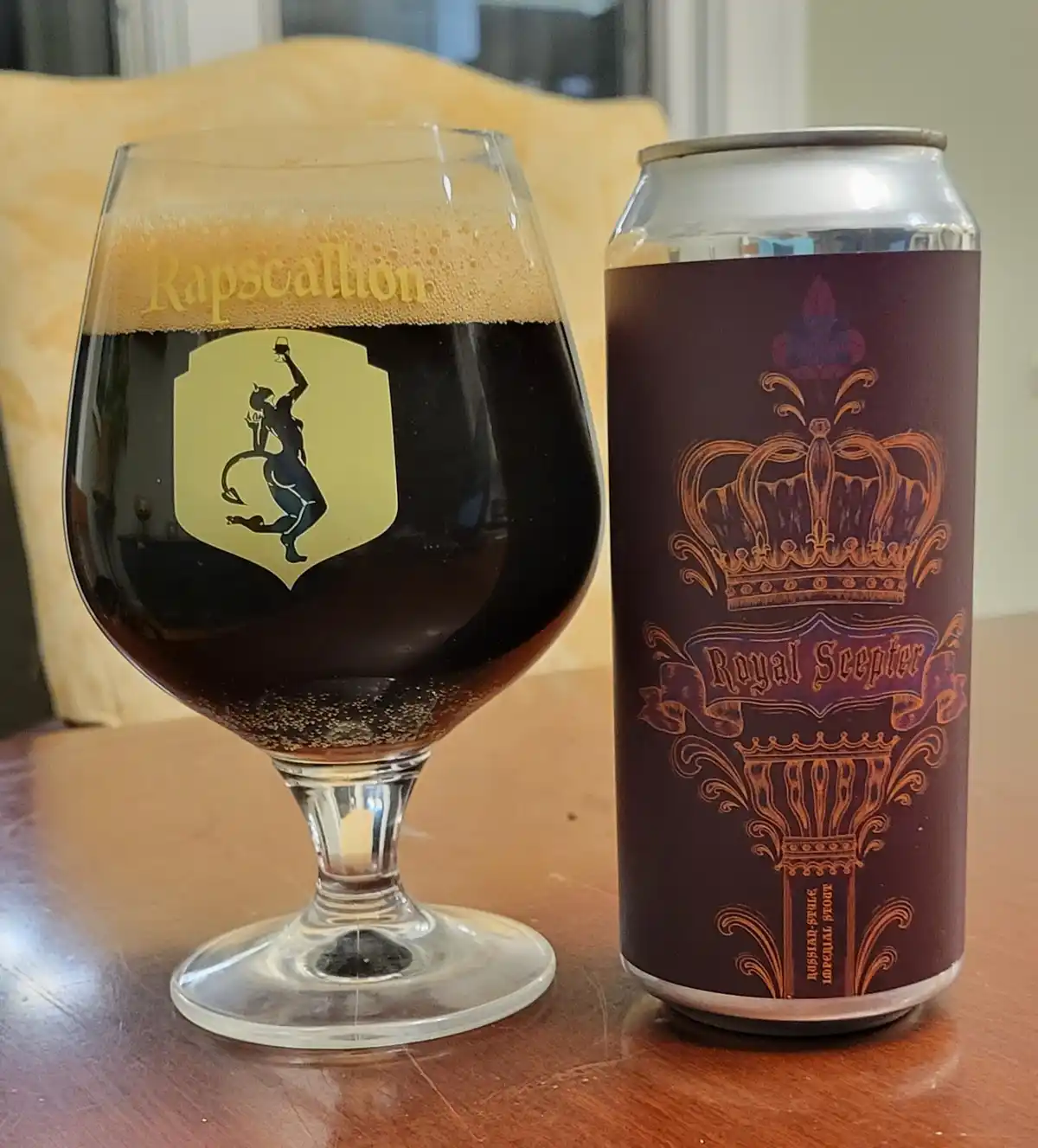 A dark beer in a stemmed glass with 'Rapscallion' etched on the foam and a devil logo, sits next to a brown can labeled…