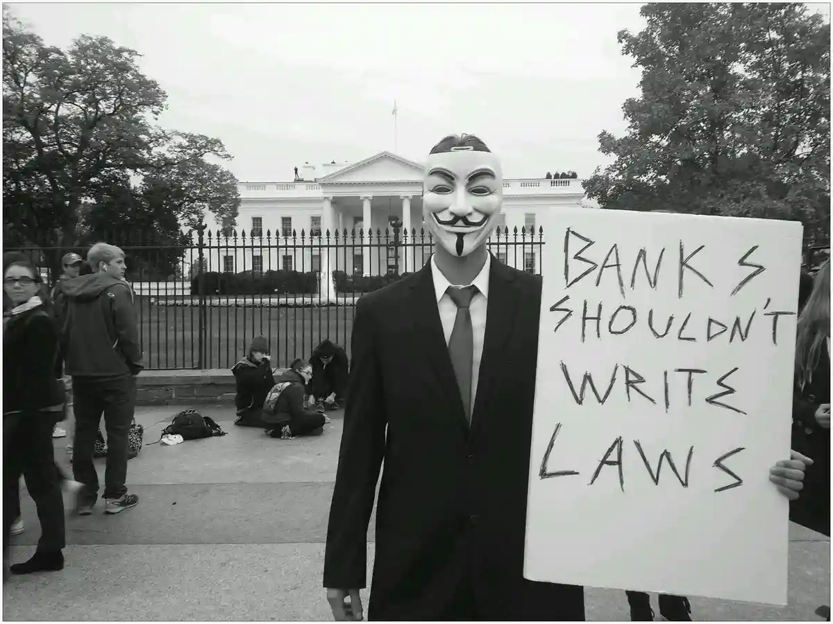 A person wearing a Guy Fawkes mask and a suit holds a handmade sign reading 'BANKS SHOULDN'T WRITE LAWS' in front of the…
