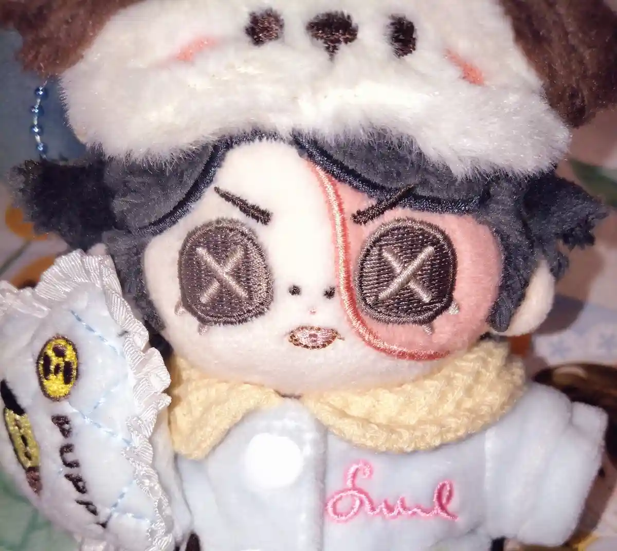 A plush doll with a split face, one side pale and the other pink, both with stitched X's for eyes, wearing a white coat with…