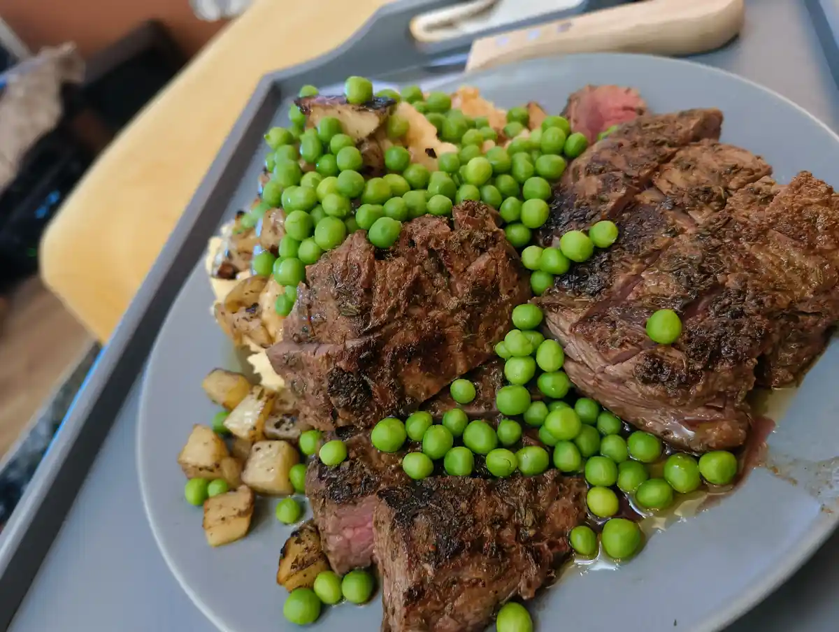 A close-up photo of a plated meal featuring sliced, herb-seasoned steak with a pink center, accompanied by roasted potatoes…