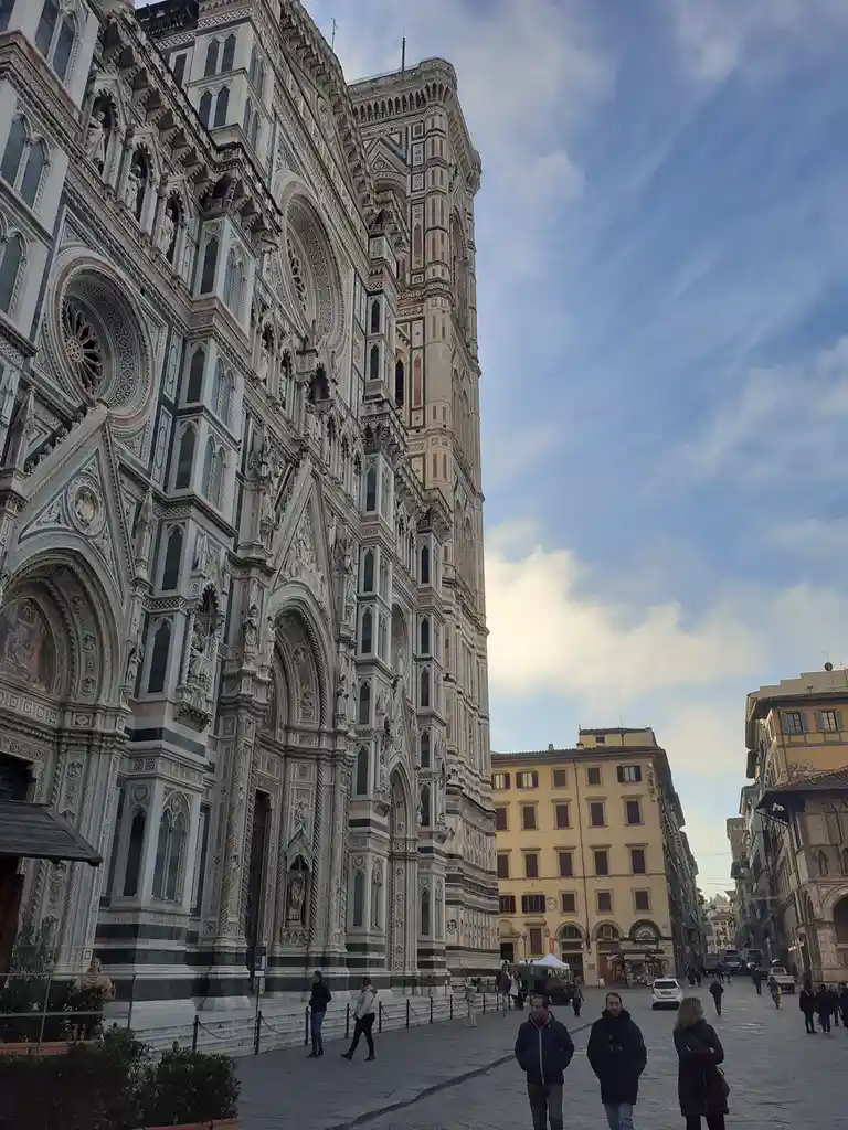 A low-angle photo of the ornate, multi-colored facade of Florence's Duomo cathedral under a partly cloudy sky, with several…