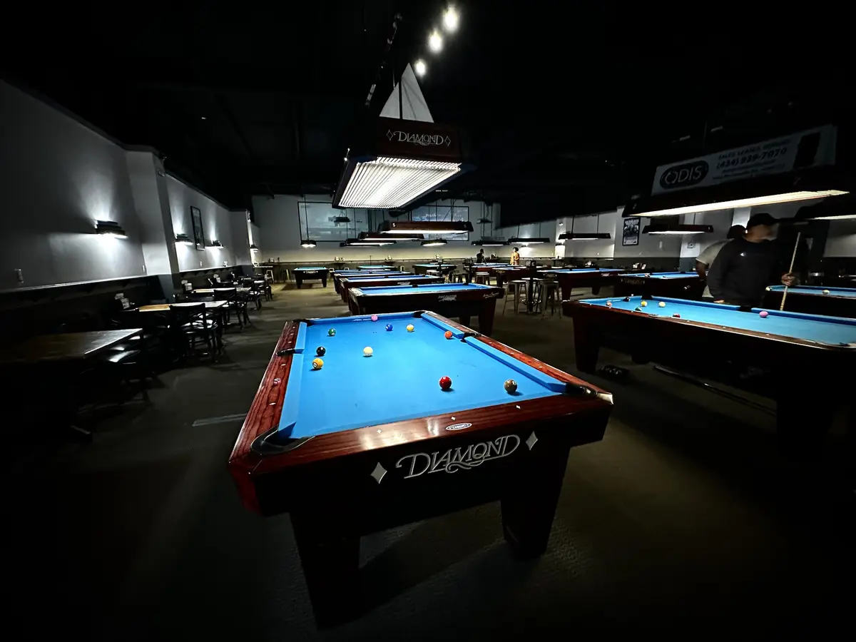A dimly lit pool hall with multiple blue-felted Diamond pool tables, some with balls scattered, and a few people playing in…