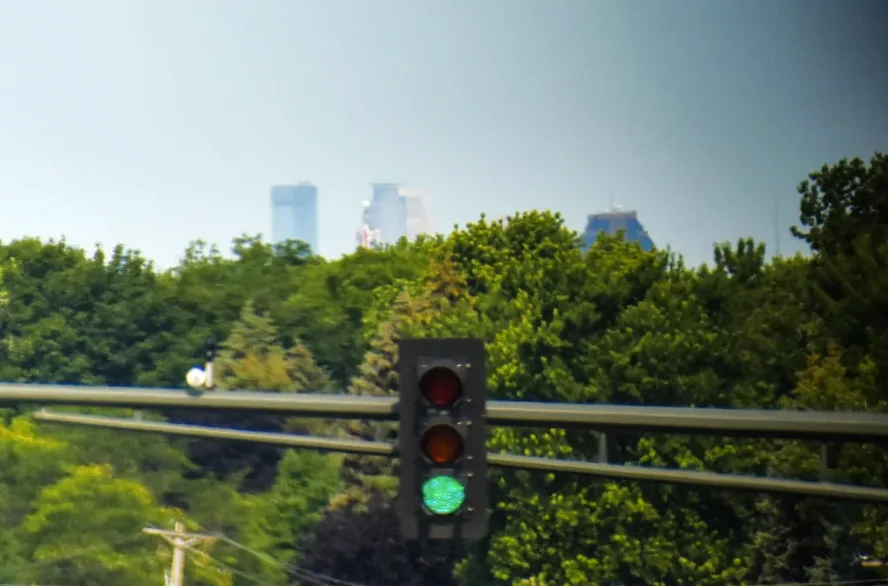 A traffic light with the green light illuminated, set against a backdrop of lush green trees and distant city skyscrapers…