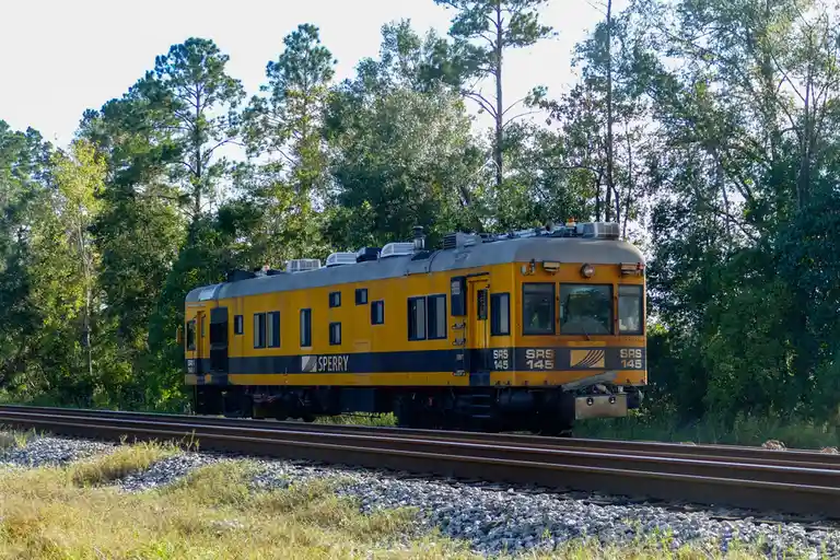 A yellow and black SRS 145 railcar, branded with 'SPERRY', travels along a track through a forested area. The overlaid text…